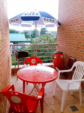 A balcony with a red table and chairs, a blue and white umbrella, and a view of greenery and buildings in the background. - Naksan Crova Condotel (Yangyang)