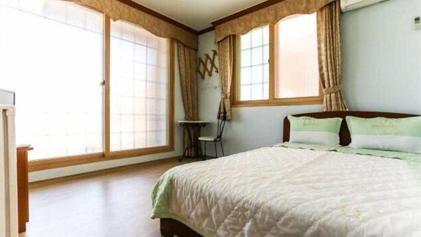 Ein Schlafzimmer mit einem Bett, einem Sessel, einem kleinen Tisch und großen Fenstern mit Vorhängen.