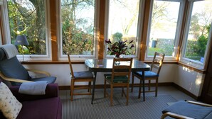 A cozy room with a purple sofa, a dining table with chairs, and large windows offering a view of greenery outside. - Calluna Cottage -  a cottage that sleeps 7 guests  in 3 bedrooms (Dalbeattie)