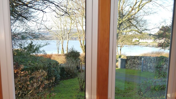 A dining area with a glass table and chairs, a vase with flowers, and a view of a garden and water body through large windows. - Calluna Cottage -  a cottage that sleeps 7 guests  in 3 bedrooms (Dalbeattie)