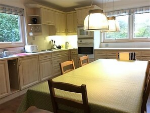 Una cocina con armarios de madera, una mesa de comedor, sillas, una lámpara colgante y una ventana con persianas.