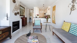 A modern living room with a fireplace, a sofa, a coffee table with magazines, and a dining area with a chair and table. - 211 De Waterkant Piazza (Cape Town)