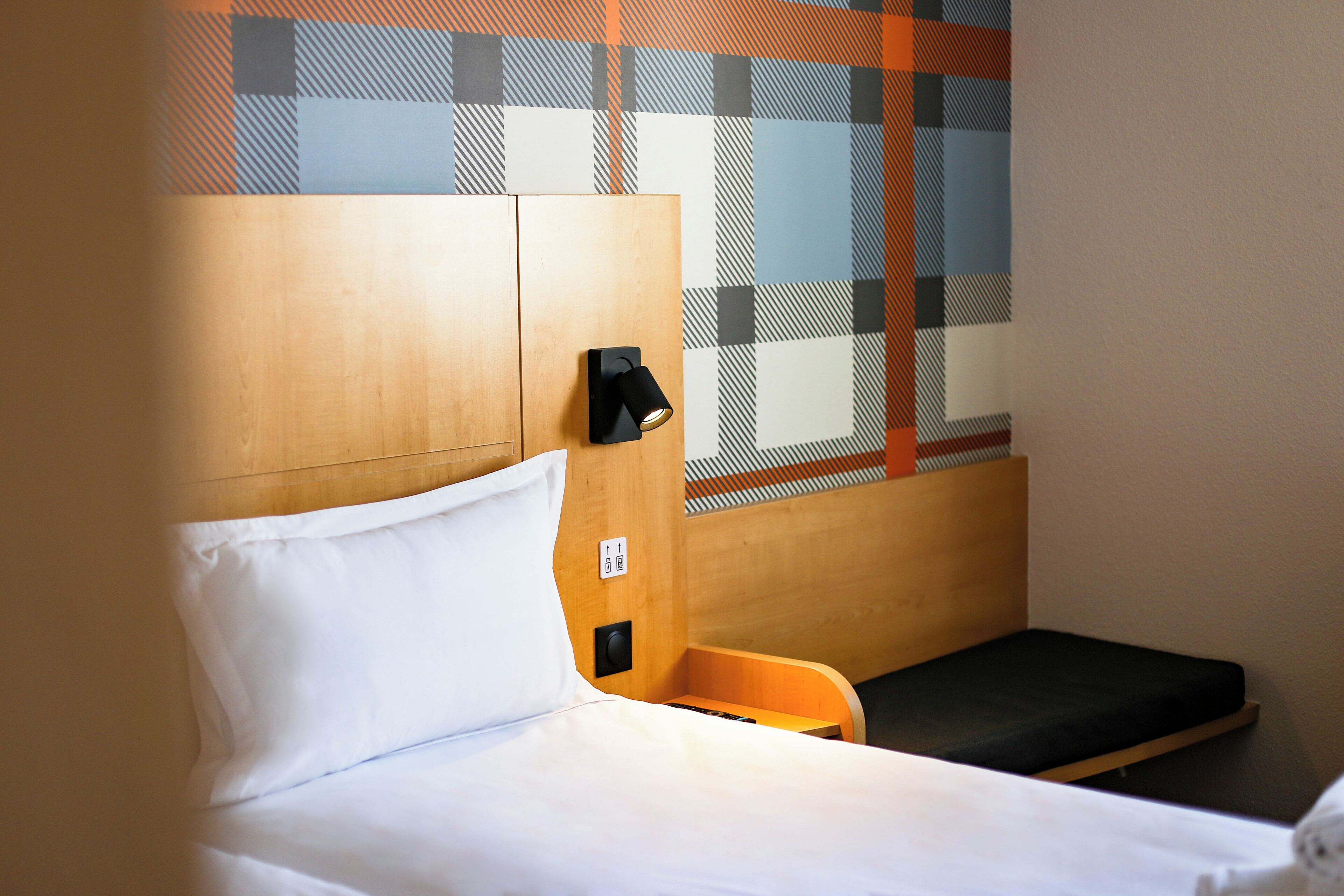 A hotel room with a bed, a small wooden bench, a wall-mounted light, and a checkered window pattern.