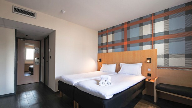 A hotel room with a large bed, a wooden headboard, a bench, and a plaid-patterned wall. - Easyhotel Nice (Nice)