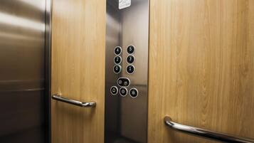 L’intérieur d’un ascenseur est doté de portes en bois, d’un panneau de commande en acier inoxydable avec des boutons permettant de sélectionner les étages, et d’un panneau indiquant que le service est assuré 24 heures sur 24, 7 jours sur 7.