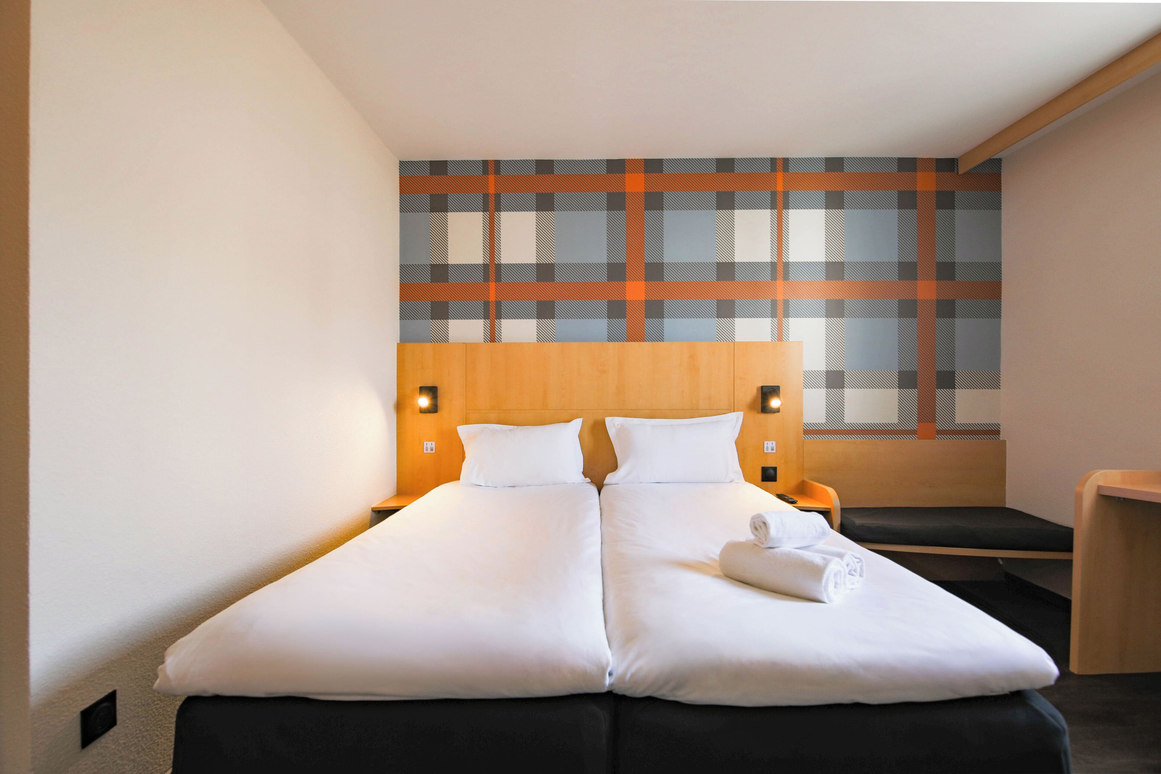 Un lit double avec du linge de lit blanc, une tête de lit en bois et un mur orné d’un motif à carreaux.
