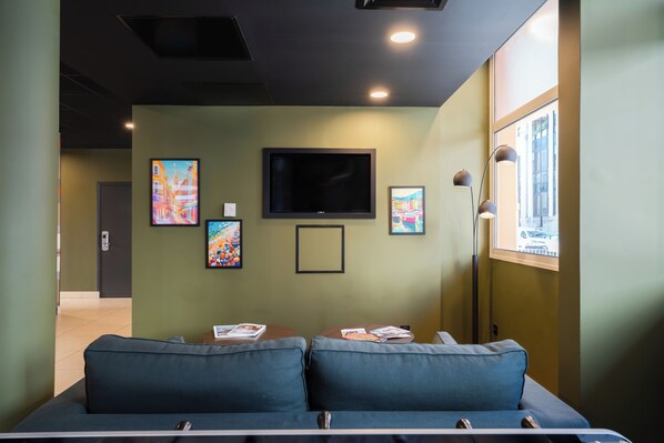 A waiting area with a flat-screen TV mounted on the wall, two blue cushioned seating areas, and framed artworks on the walls. - Easyhotel Nice (Nice)