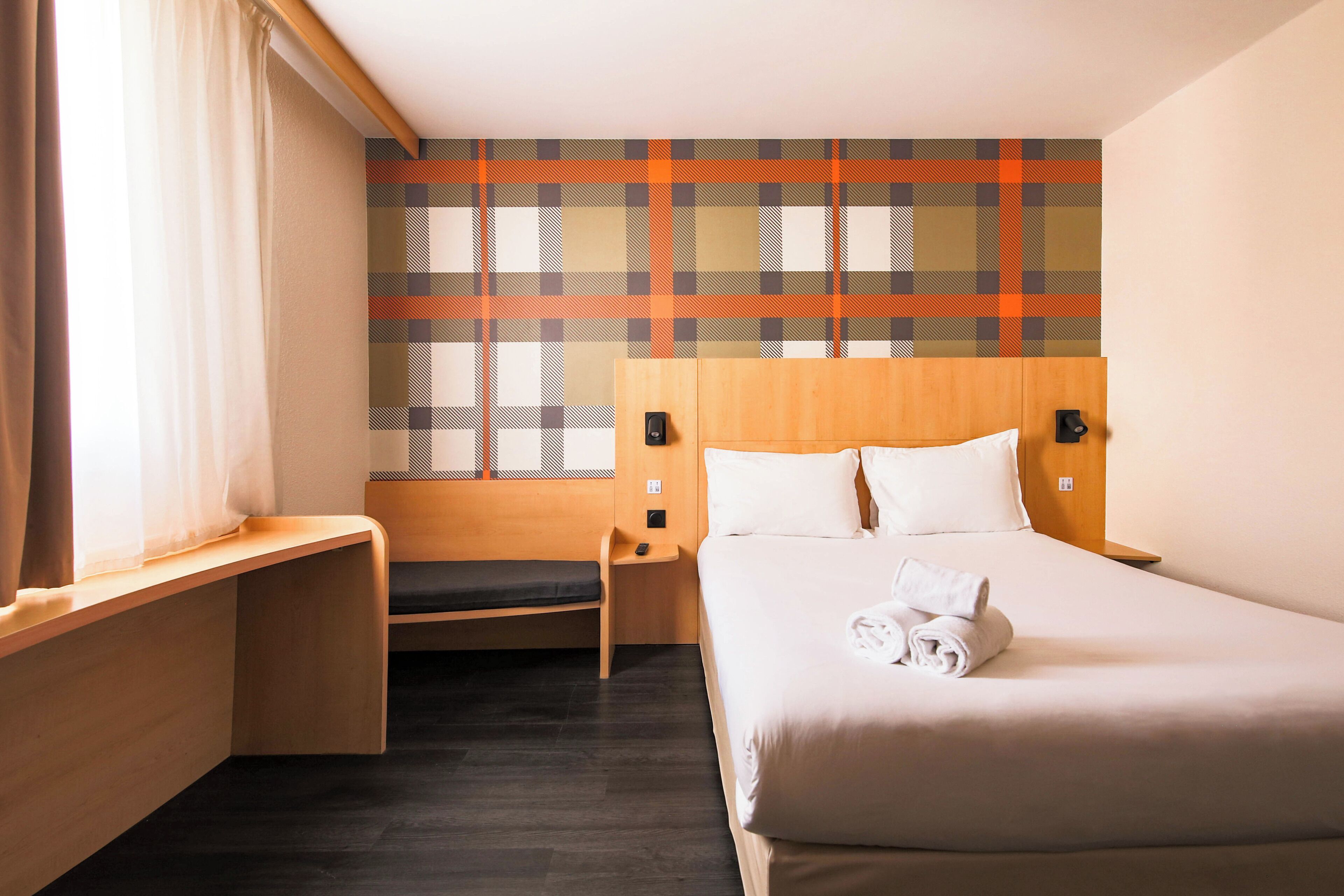 A hotel room with a bed, a bench, a nightstand, and a wall with a plaid pattern.