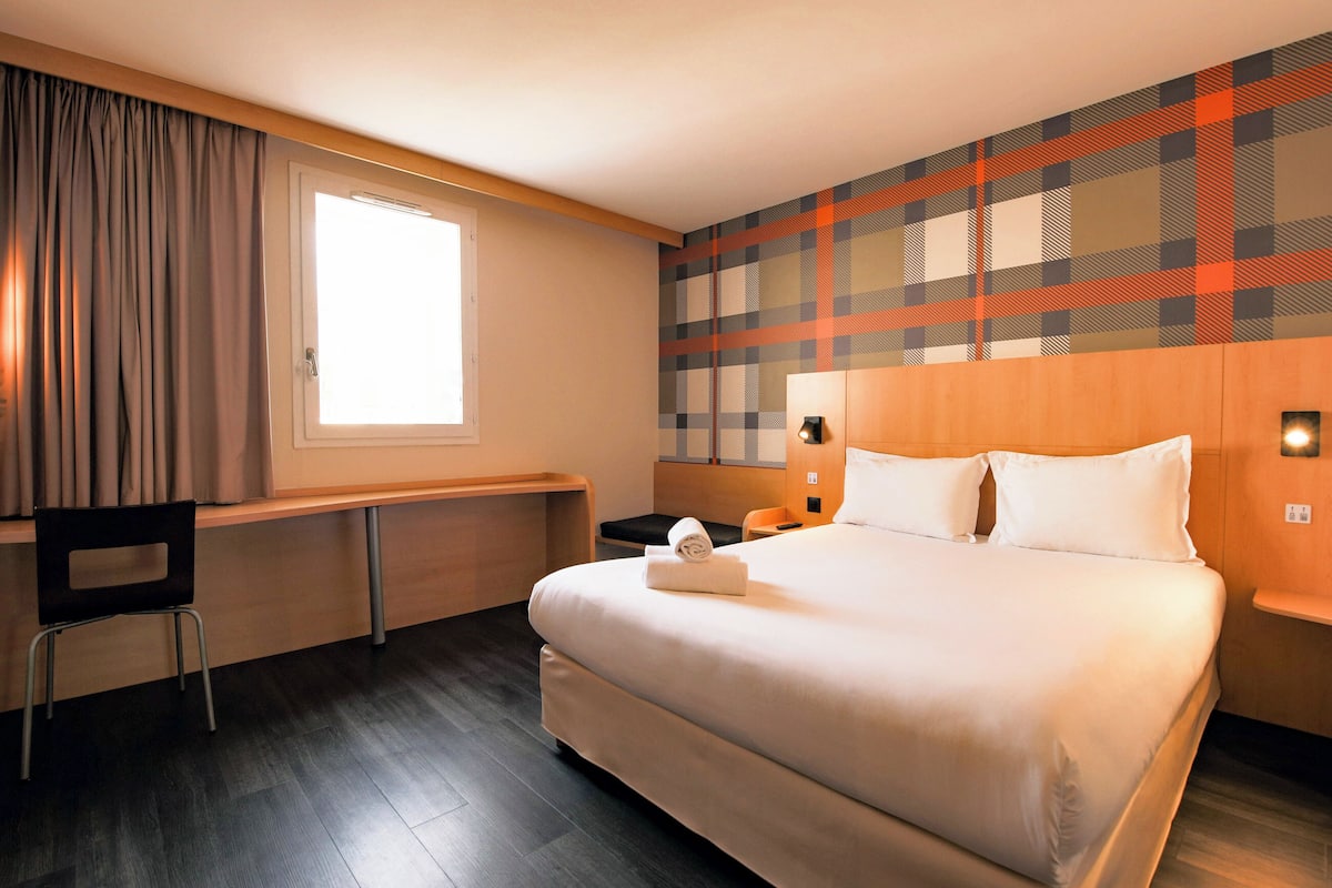 A hotel room with a large bed, a desk, a chair, a window with curtains, and a plaid-patterned wall.