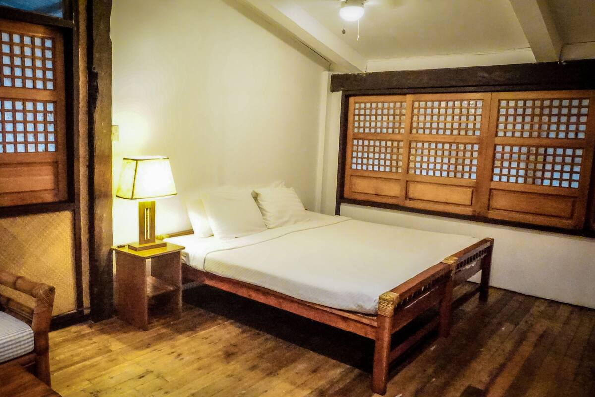 A traditional room with a wooden bed, a nightstand with a lamp, a wooden chair, and a window with wooden lattice.