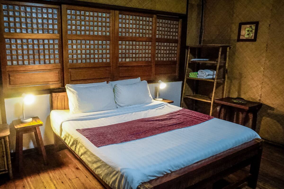 A traditional wooden bed with white linens and a red throw blanket, flanked by nightstands with lamps, in a room with a wooden headboard and a shelf with towels.
