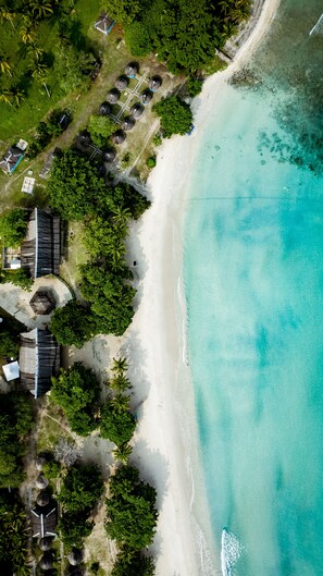 An aerial view of a beach resort with a sandy shoreline, clear turquoise waters, and several structures including a large building and smaller huts. - Vista del Mar by Cocotel (Zamboanga)