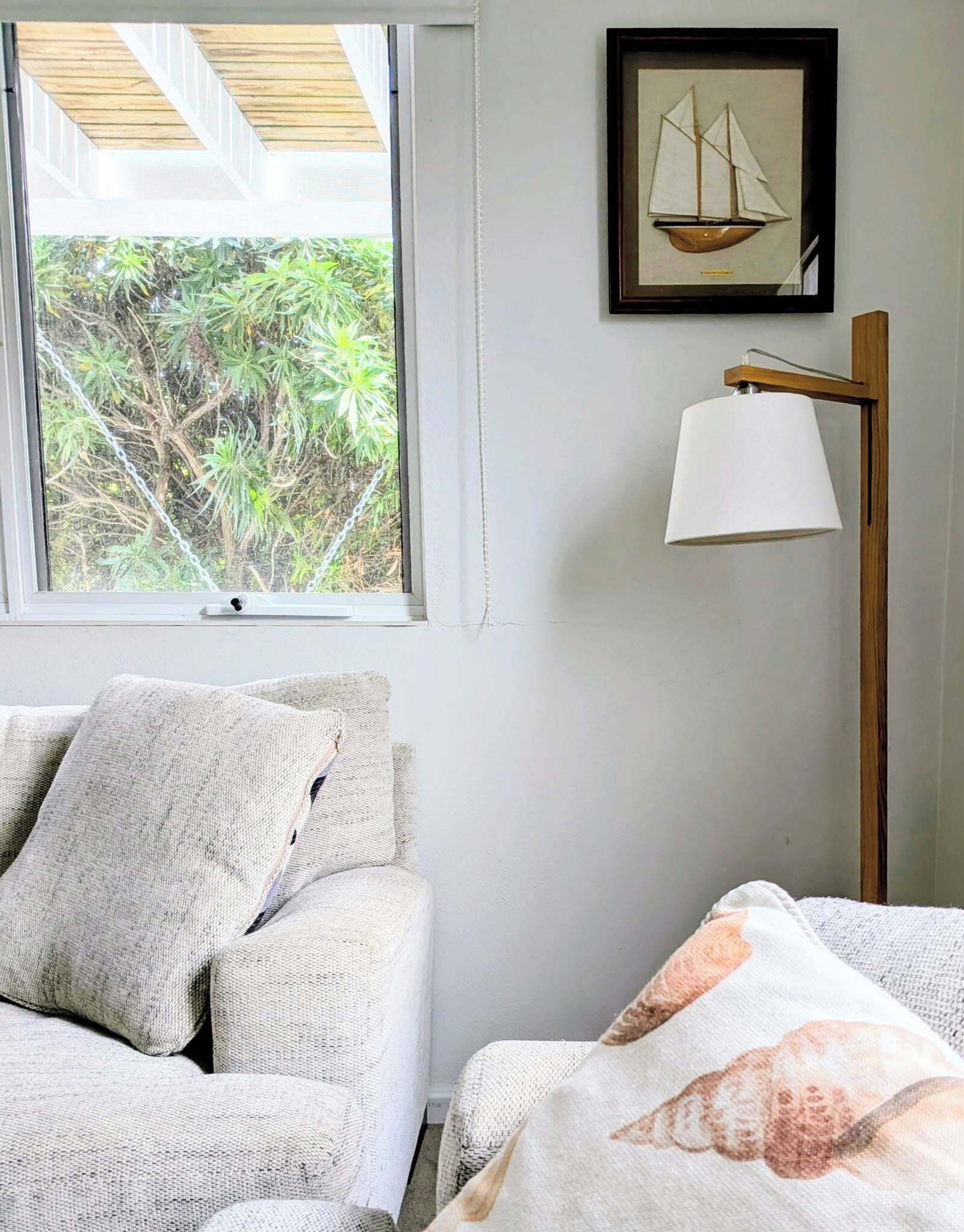 A room with a bed, a window with a view of greenery, a framed picture on the wall, and a floor lamp.