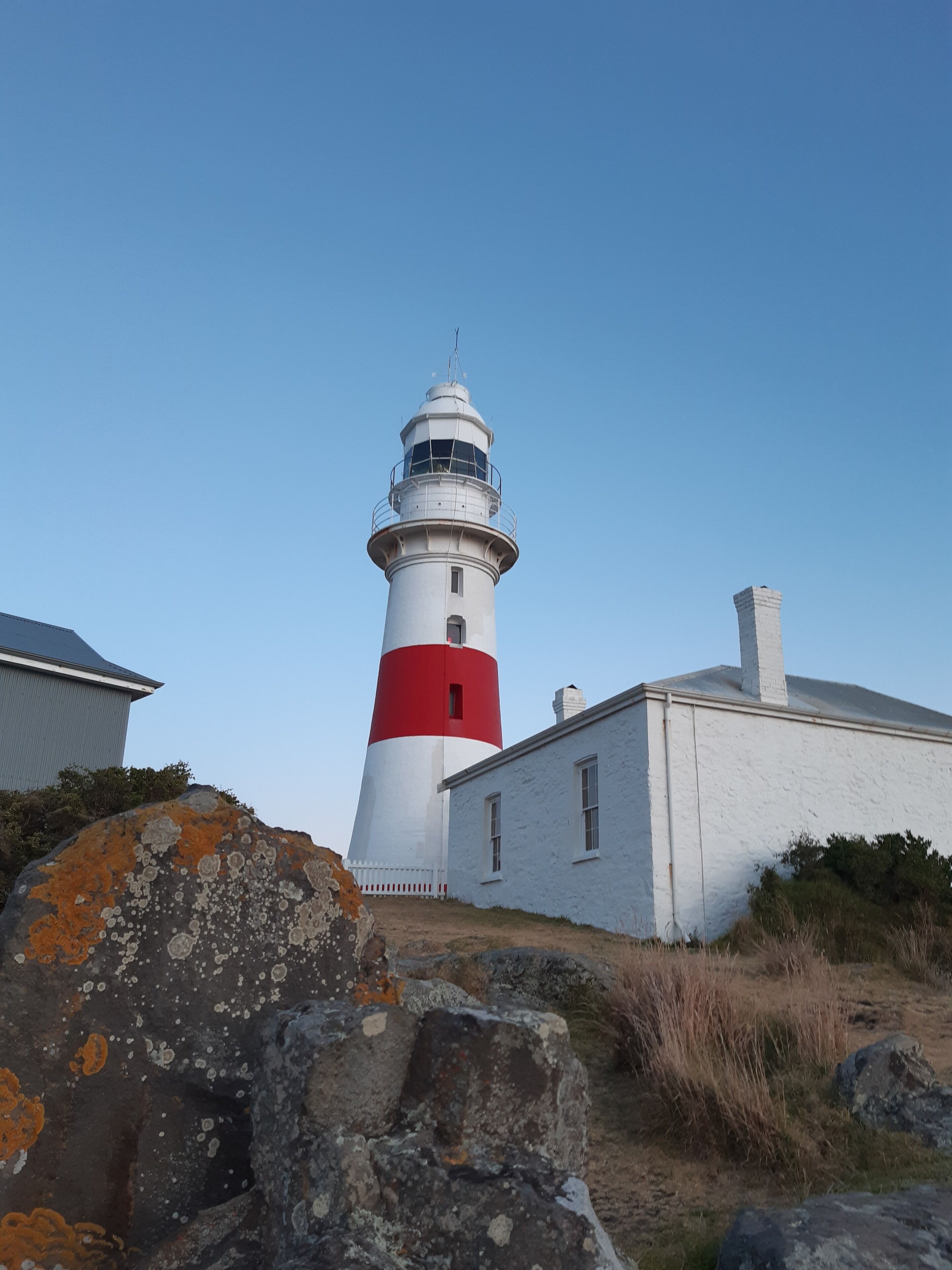 A lighthouse with a red and white structure, a white building adjacent to it, and surrounding rocks and vegetation.