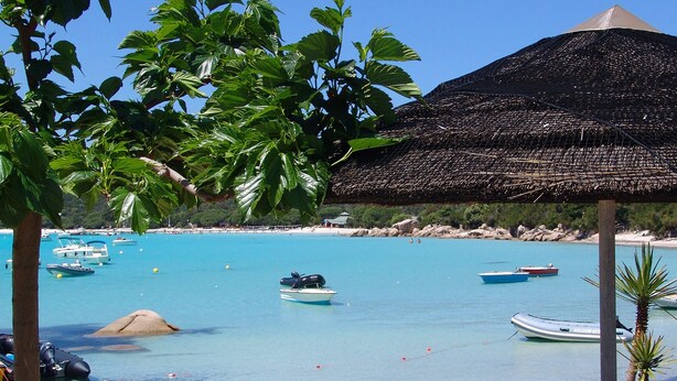 Una playa con agua turquesa cristalina, varios barcos y una estructura de techo de paja.
