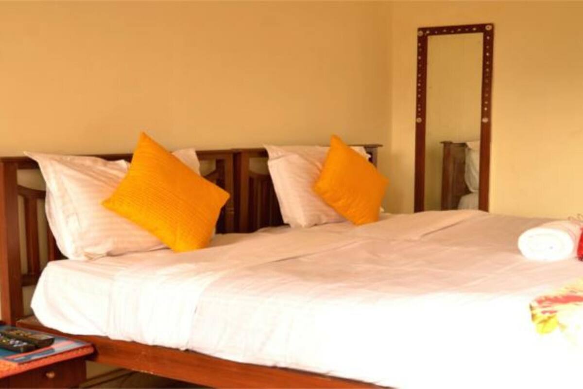 A bed with white bedding and two orange pillows, a bedside table with a remote control, and a mirror on the wall.