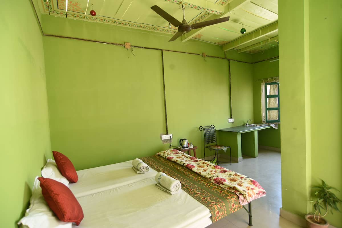 A room with a bed, a ceiling fan, a small table with a kettle, a chair, and a window.