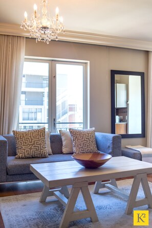 A modern living room with a sofa, a wooden coffee table, a chandelier, and a large window with curtains. - Unit 613 Cape Royale Apartments (Cape Town)
