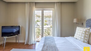 A hotel room with a bed, a TV on a stand, a window with curtains, and a view of buildings outside. - Unit 613 Cape Royale Apartments (Cape Town)