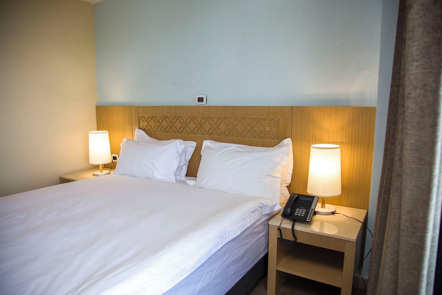 A hotel room with a neatly made bed, two bedside lamps, a bedside phone, and a wooden headboard.