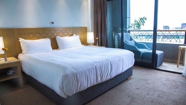 A hotel room with a large bed, a nightstand, a chair, and a view of the city through a glass door.