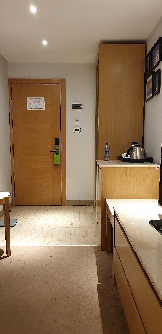 A room with a wooden door, a small kitchenette with a kettle, a water bottle, and a framed picture on the wall.