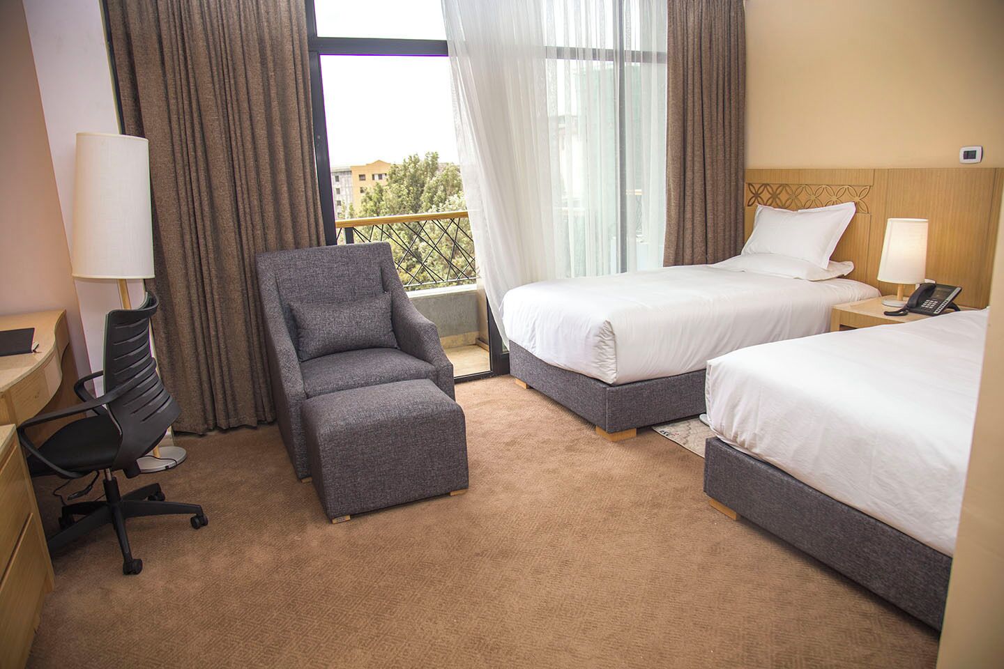 A hotel room with two beds, a desk, a chair, and a balcony with a view.