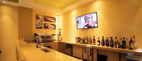 Uma área de bar com uma máquina de café, uma televisão fixada na parede e diversas garrafas de bebidas alcoólicas.