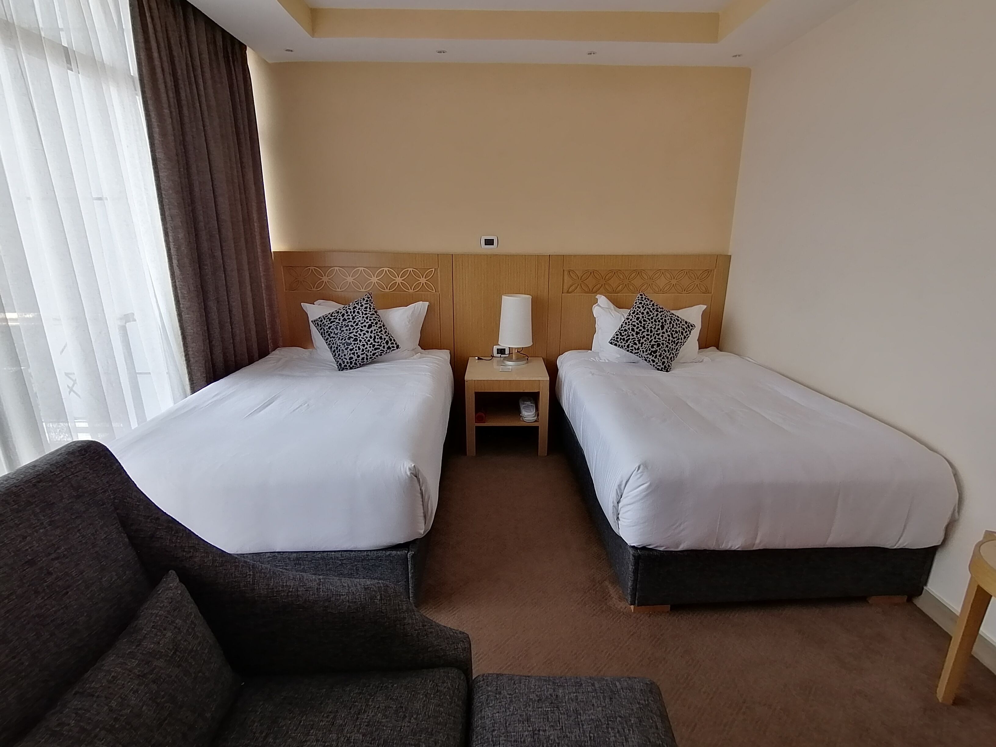 A hotel room with two beds, a nightstand, a lamp, and a sofa.