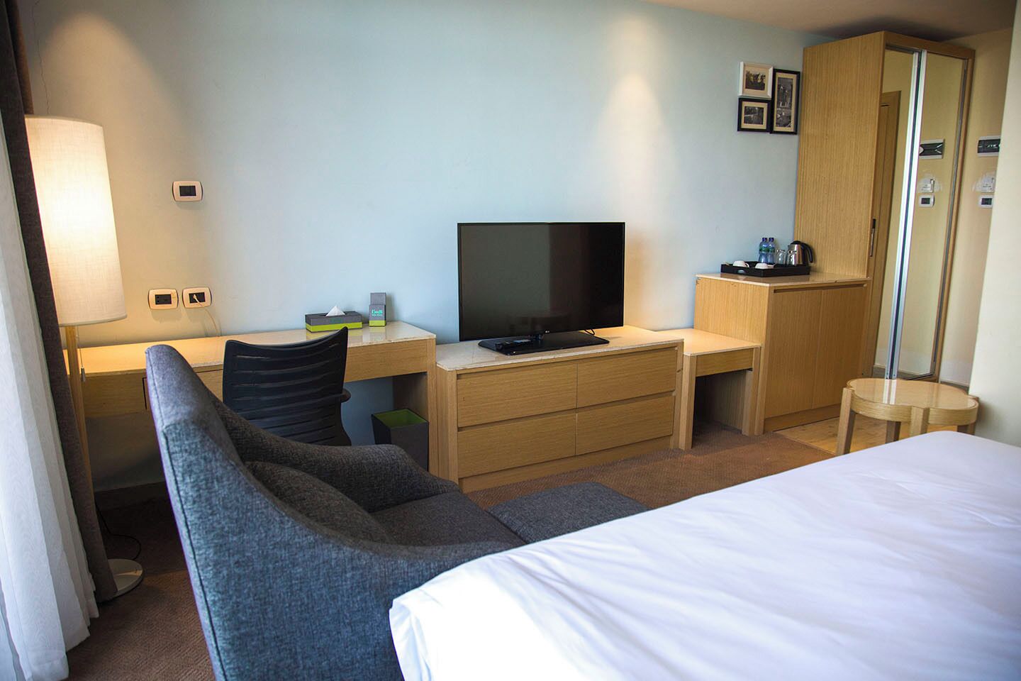 A hotel room with a bed, a desk with a television, a chair, a wardrobe, and a bedside table with a lamp.