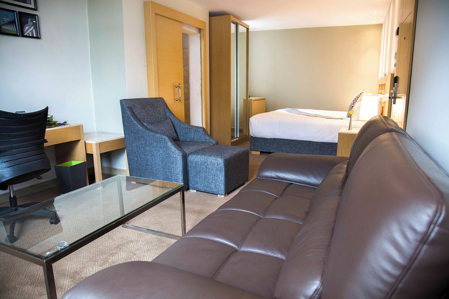 A hotel room with a leather sofa, a grey armchair, a glass coffee table, a bed with a white duvet, and a bedside table with a lamp.
