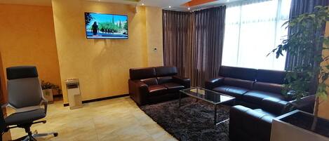 A modern lobby with a large flat-screen TV, leather sofas, a glass coffee table, and a potted plant.