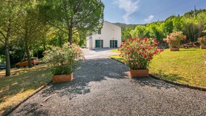 A gravel driveway leading to a white building with green shutters, surrounded by lush greenery and flowering plants. - Lovely apartment in villa for 10 guests with pool, TV and pets allowed (Rosignano Marittimo)