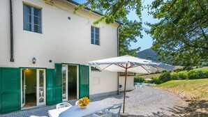 A white building with green shutters, a gravel patio, a table with chairs, and a large umbrella. - Lovely apartment in villa for 10 guests with pool, TV and pets allowed (Rosignano Marittimo)