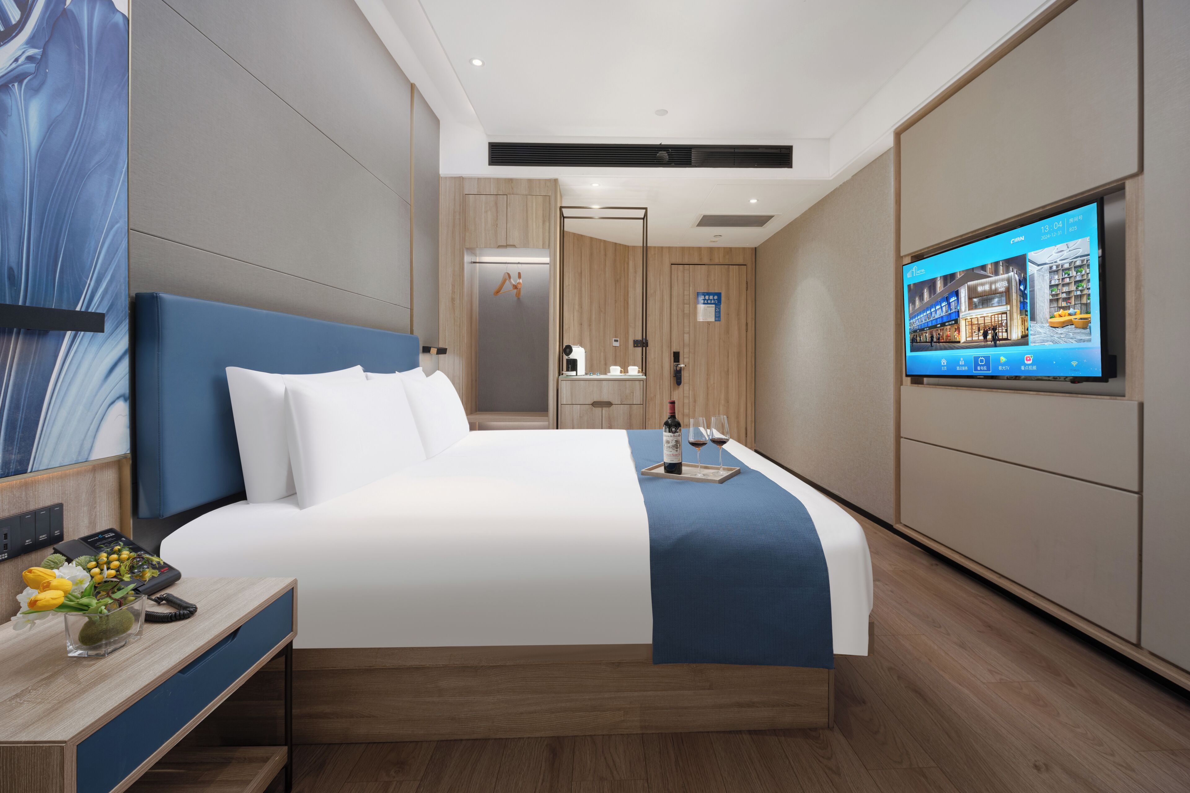 A modern hotel room with a large bed, a TV on the wall, a desk with a phone and flowers, and a bathroom visible through a glass door.