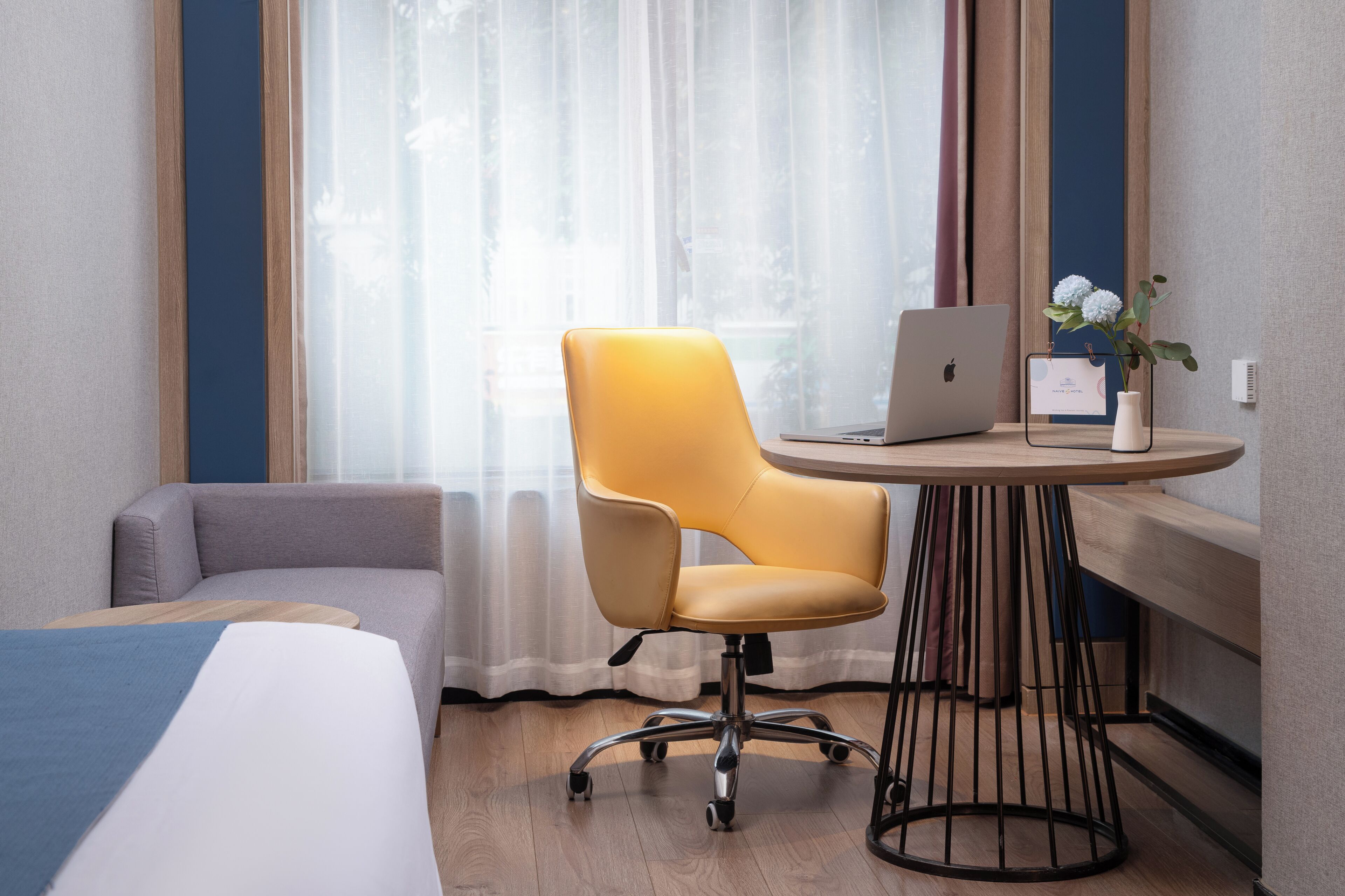 Een hotelkamer met een bed, een bureau met een laptop, een stoel en een vaas met bloemen.
