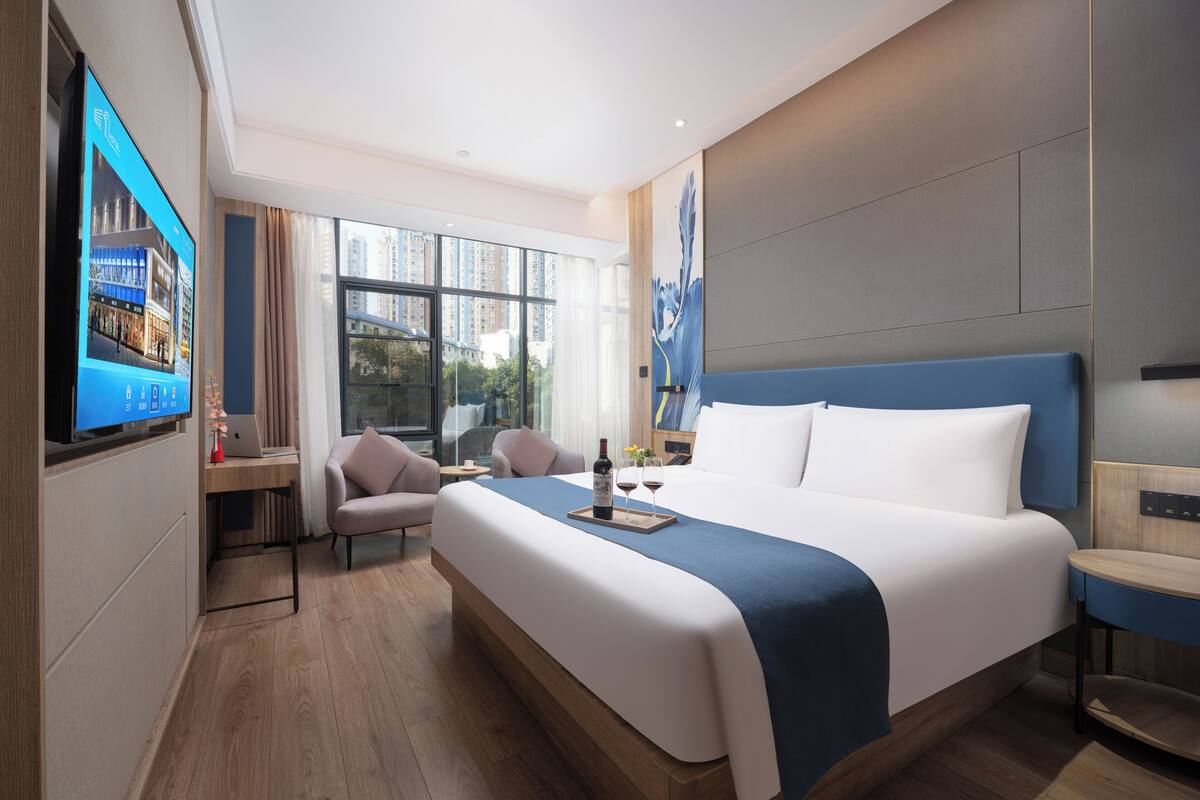 A modern hotel room with a large bed, a desk with a laptop, a TV, and a view of the city.