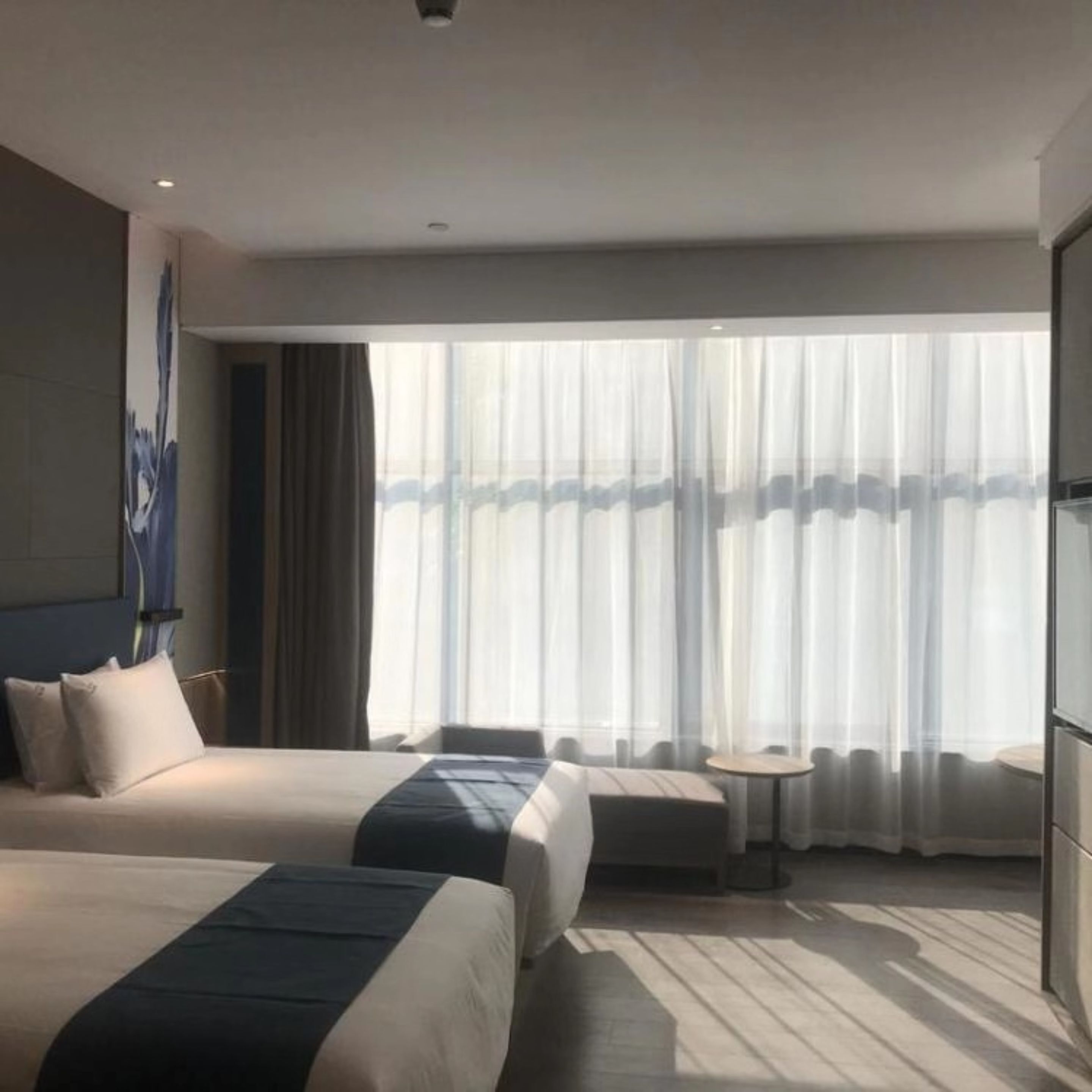 A hotel room with two beds, a small table, and a large window with sheer curtains.
