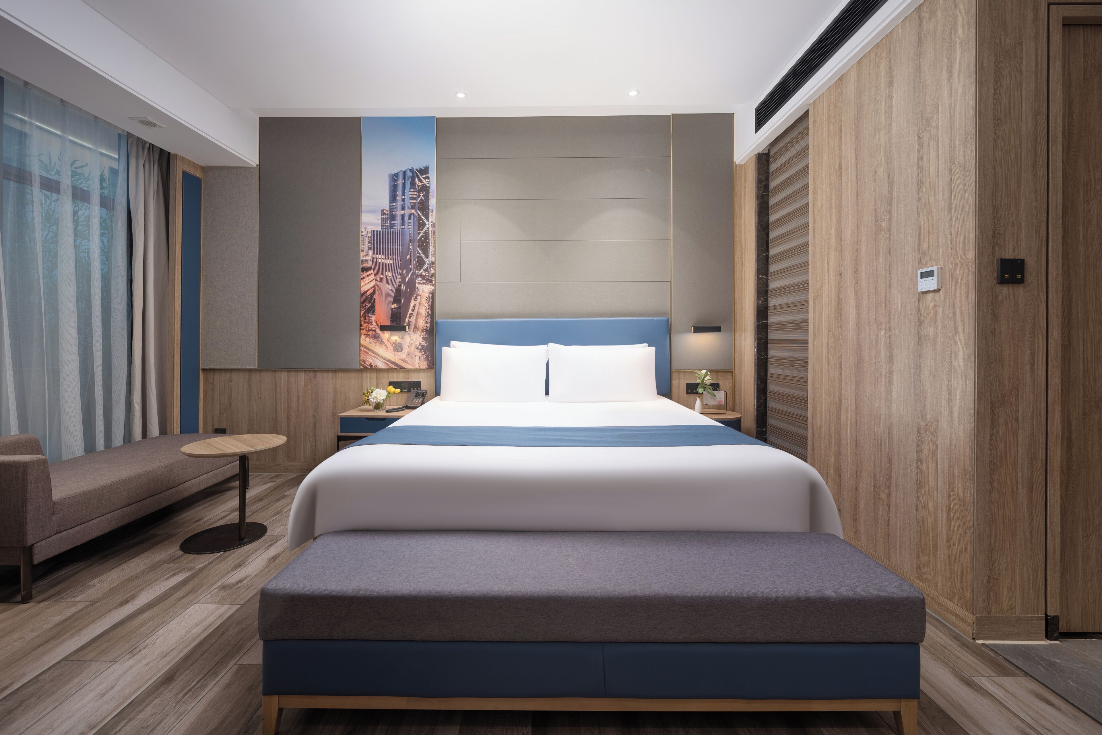 A modern hotel room with a large bed, a seating area, a small table, and a cityscape view.