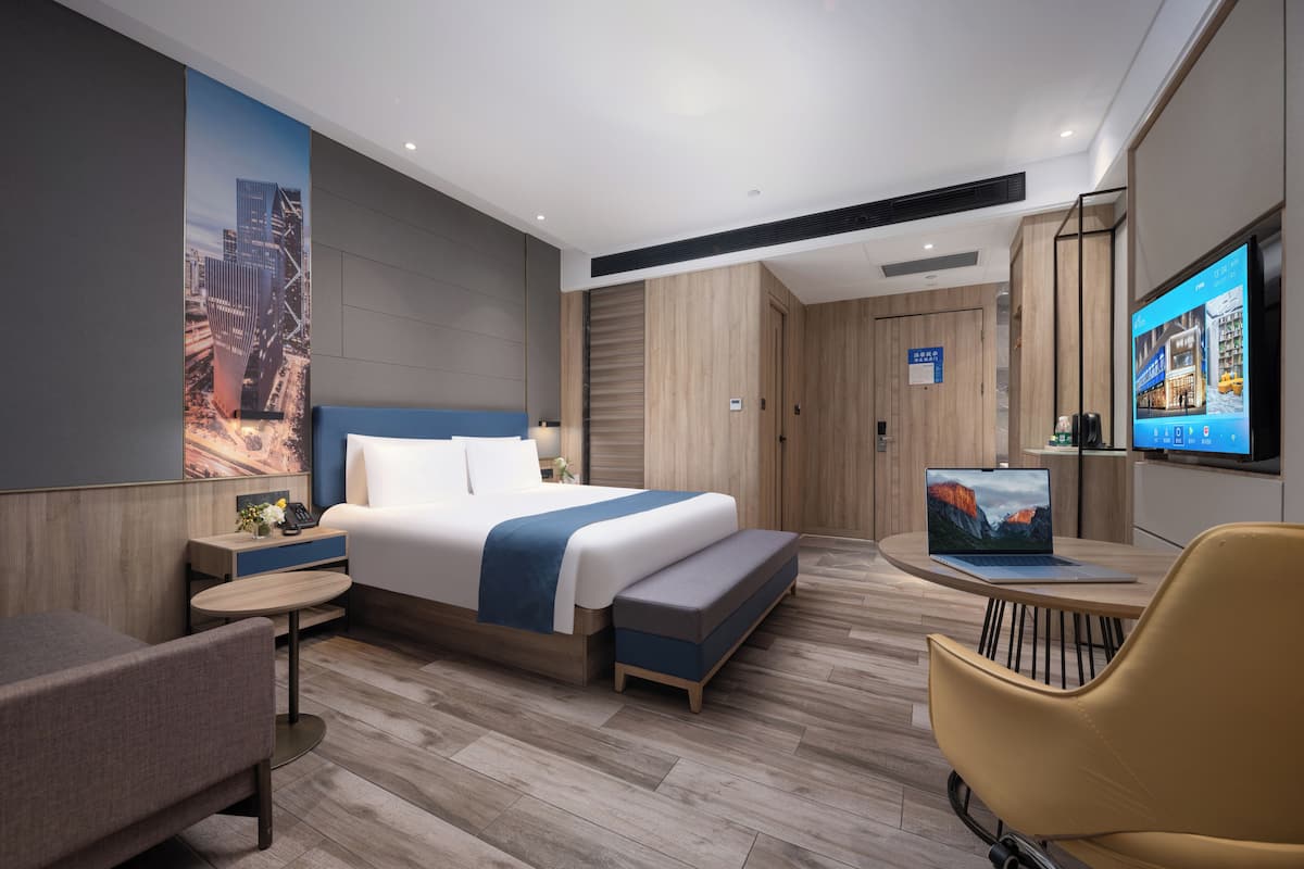 A modern hotel room with a large bed, a desk with a laptop, a TV, and a seating area with a chair.