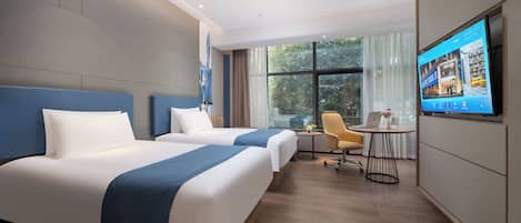 A hotel room with two beds, a desk, a chair, a TV, and a window with a view of greenery.
