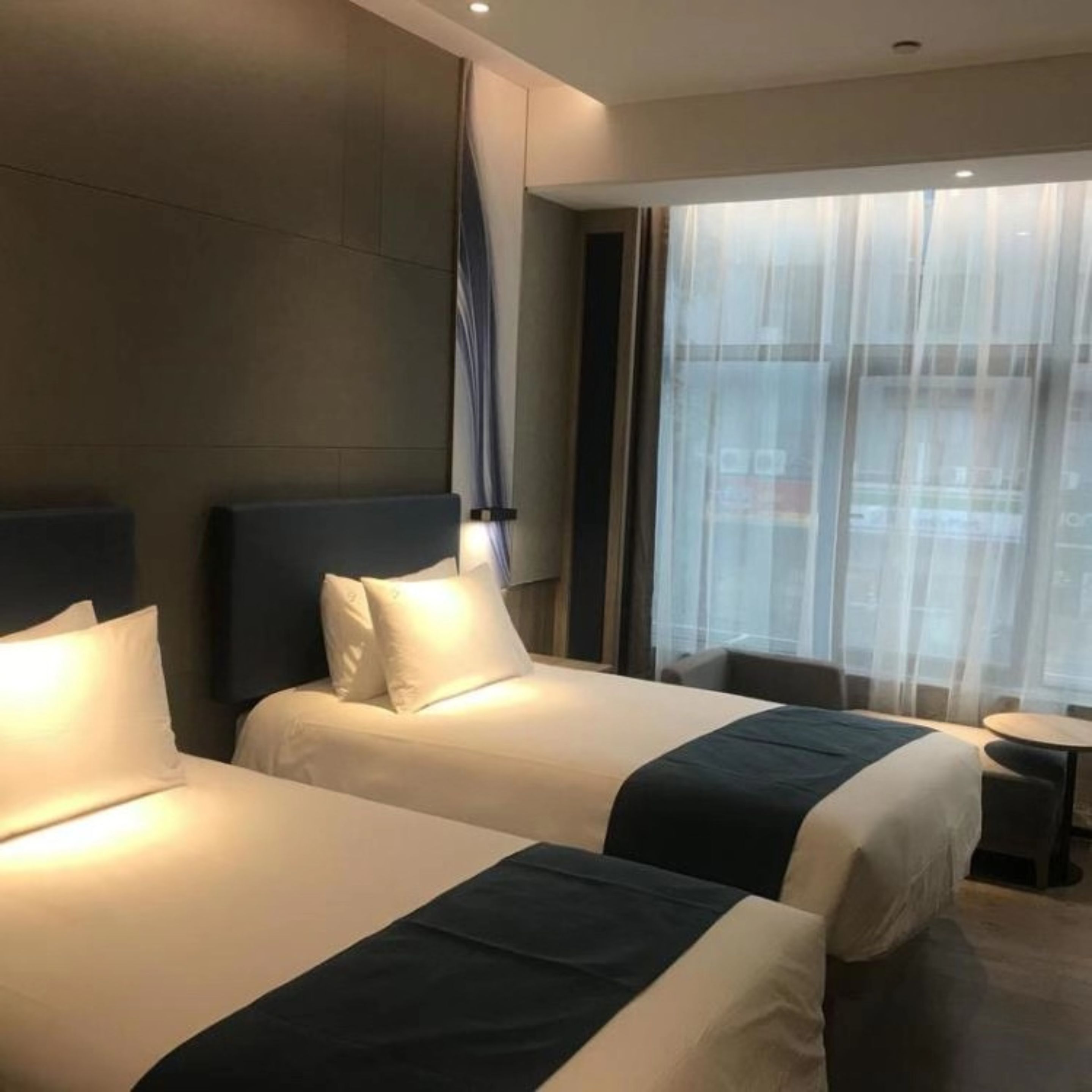 A hotel room with two beds, a small table, and a view of the city through sheer curtains.