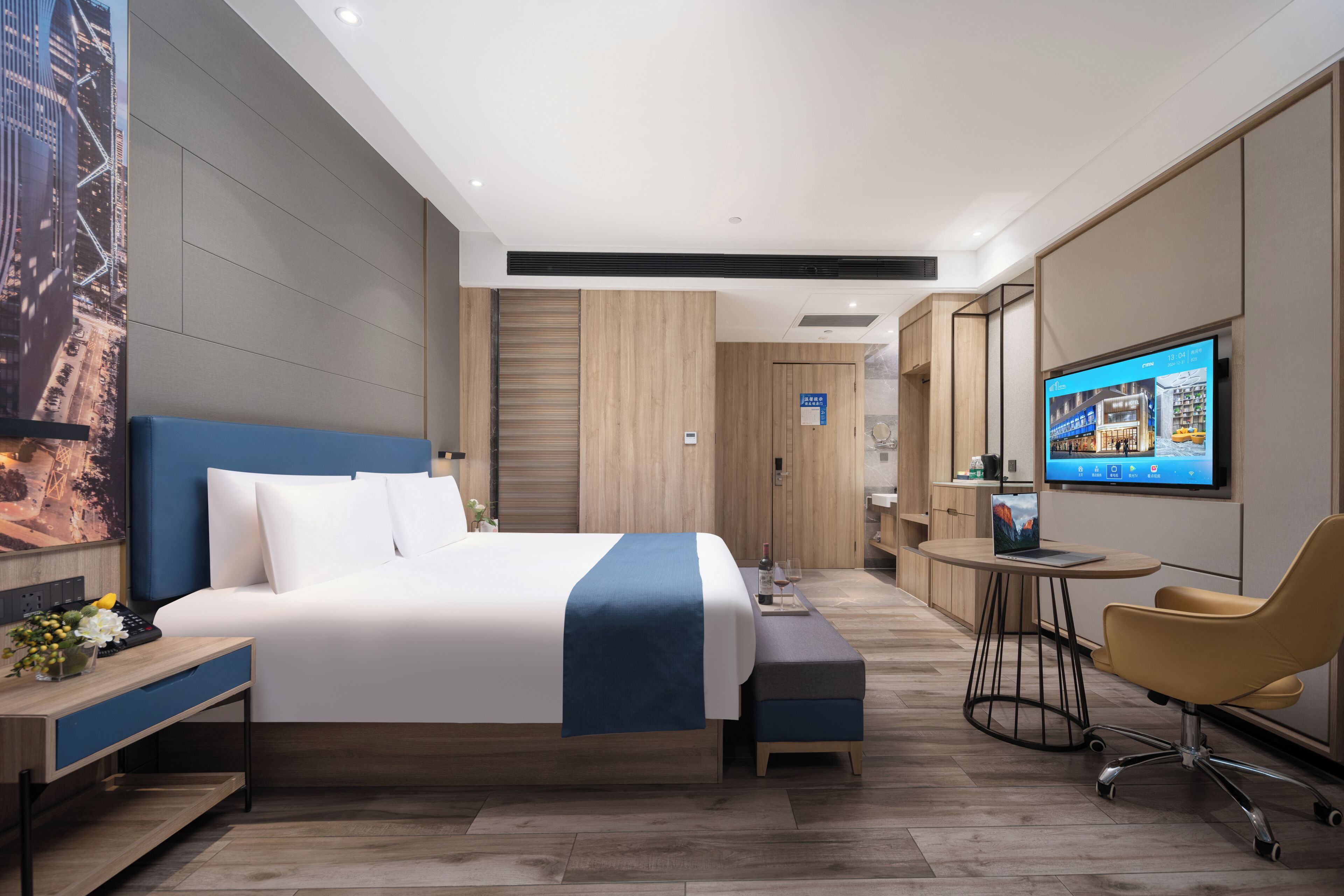 A modern hotel room with a large bed, a desk with a laptop, a TV, and a seating area with a chair.