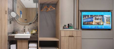 Une salle de bain moderne avec un meuble-lavabo en bois, un miroir rond, un lavabo encastré et un téléviseur diffusant une image de l’intérieur d’un hôtel.