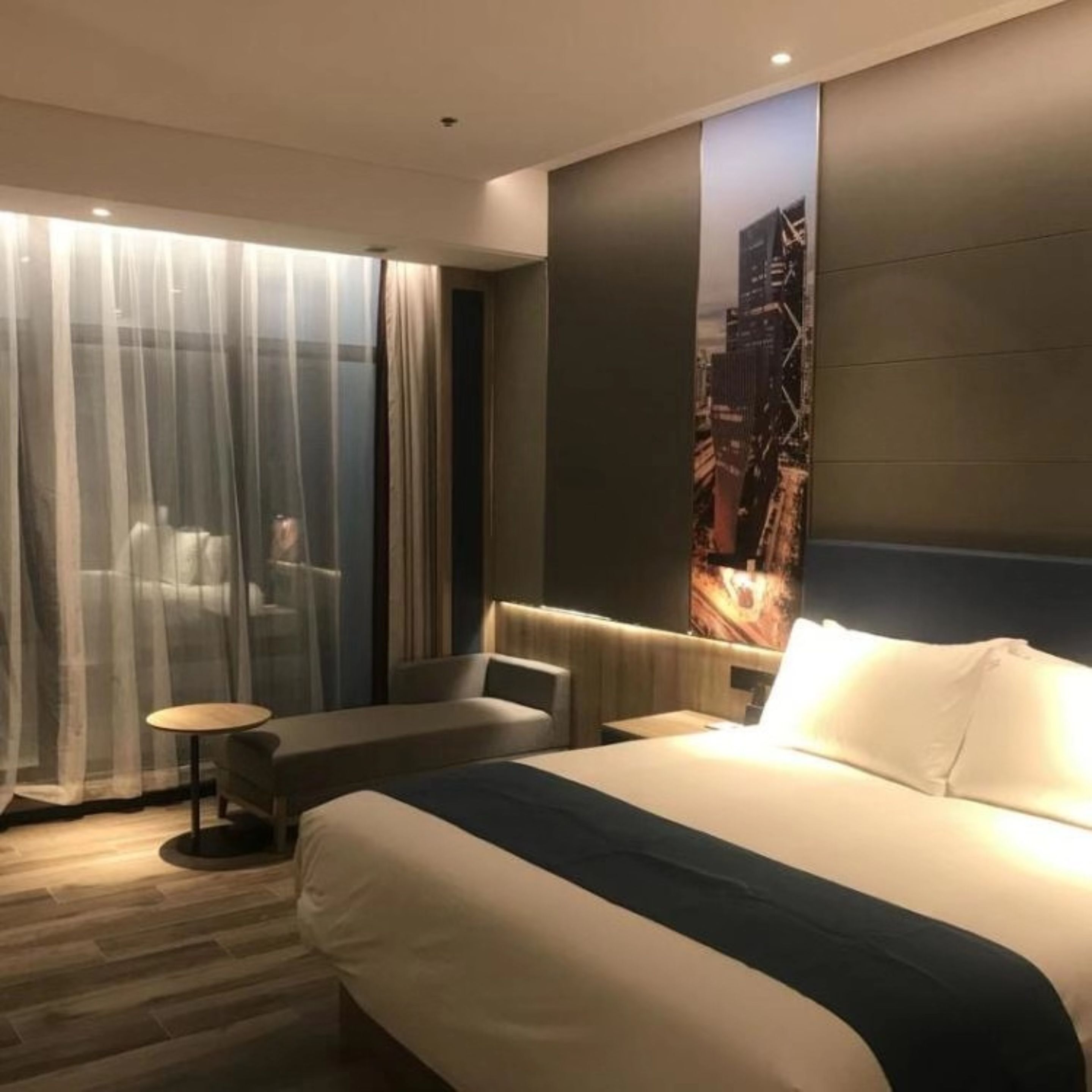 A modern hotel room with a large bed, a seating area with a small table, and a cityscape view on the wall.