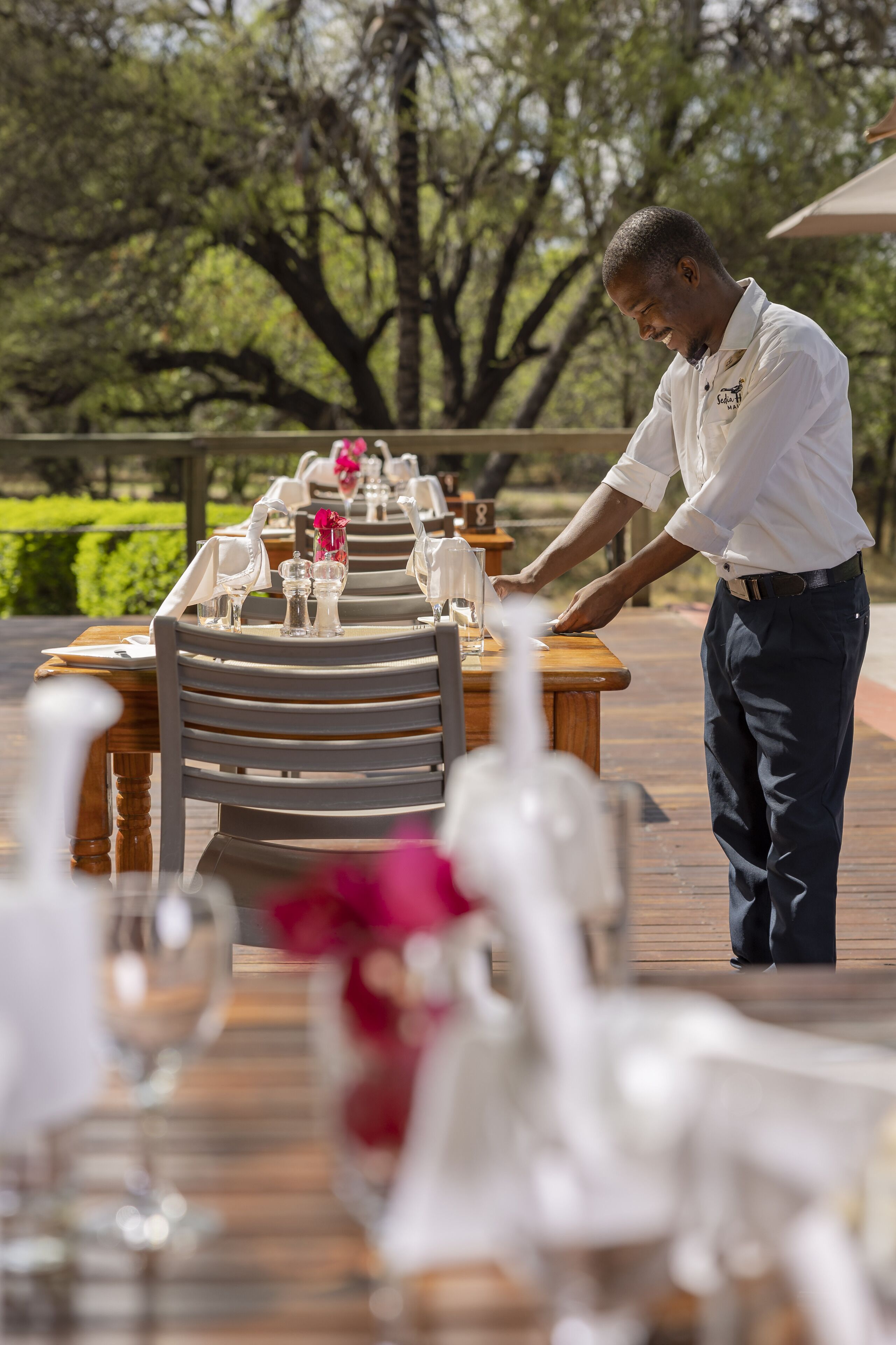 A person setting up a table outdoors with a wooden deck and greenery in the background.