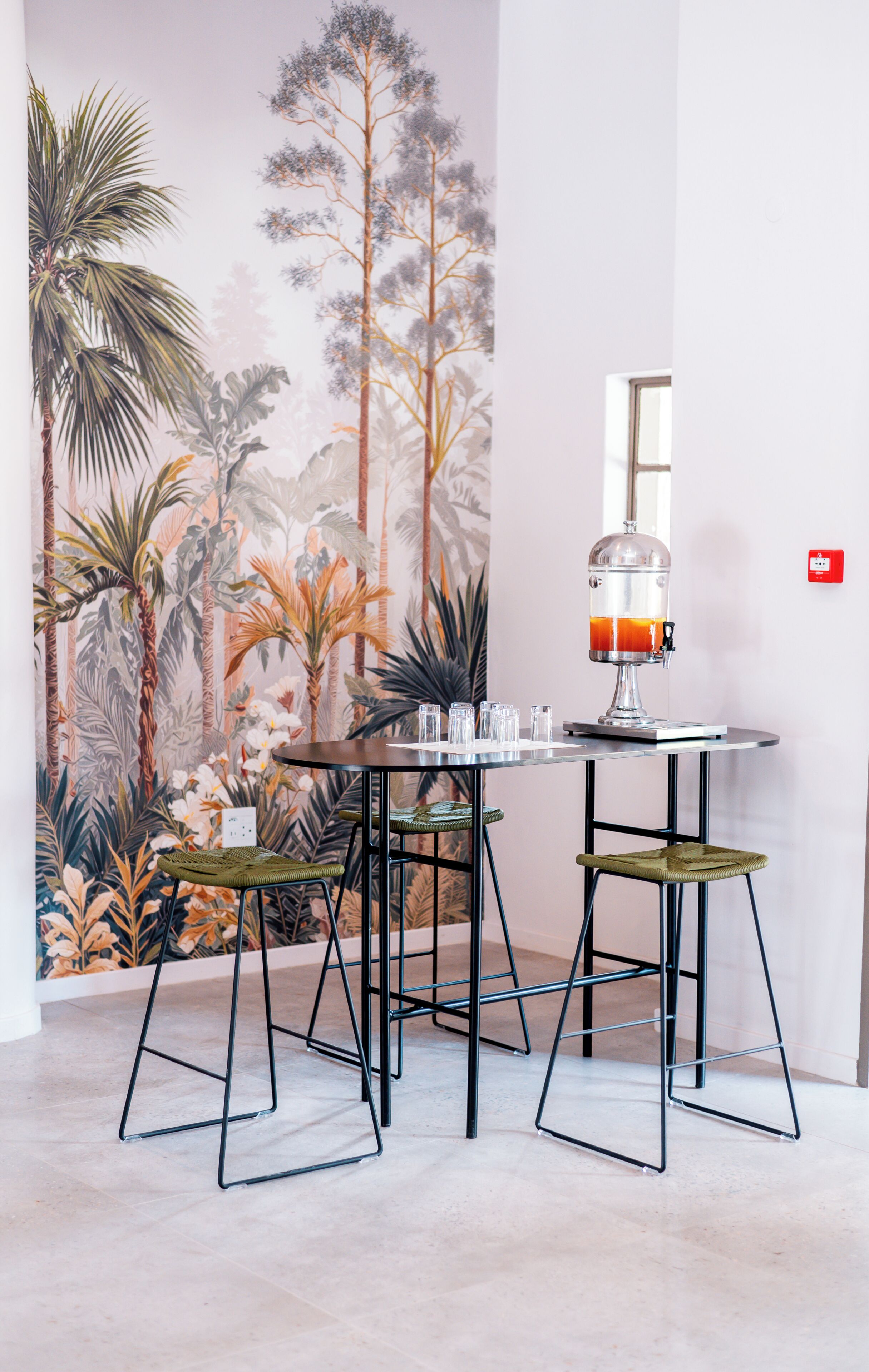 Een kamer met een muurschildering van tropische begroeiing, een tafel met een glazen blad en twee barkrukken.