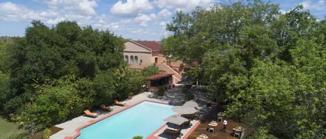 A swimming pool with lounge chairs and umbrellas, surrounded by trees and a building with a red roof.