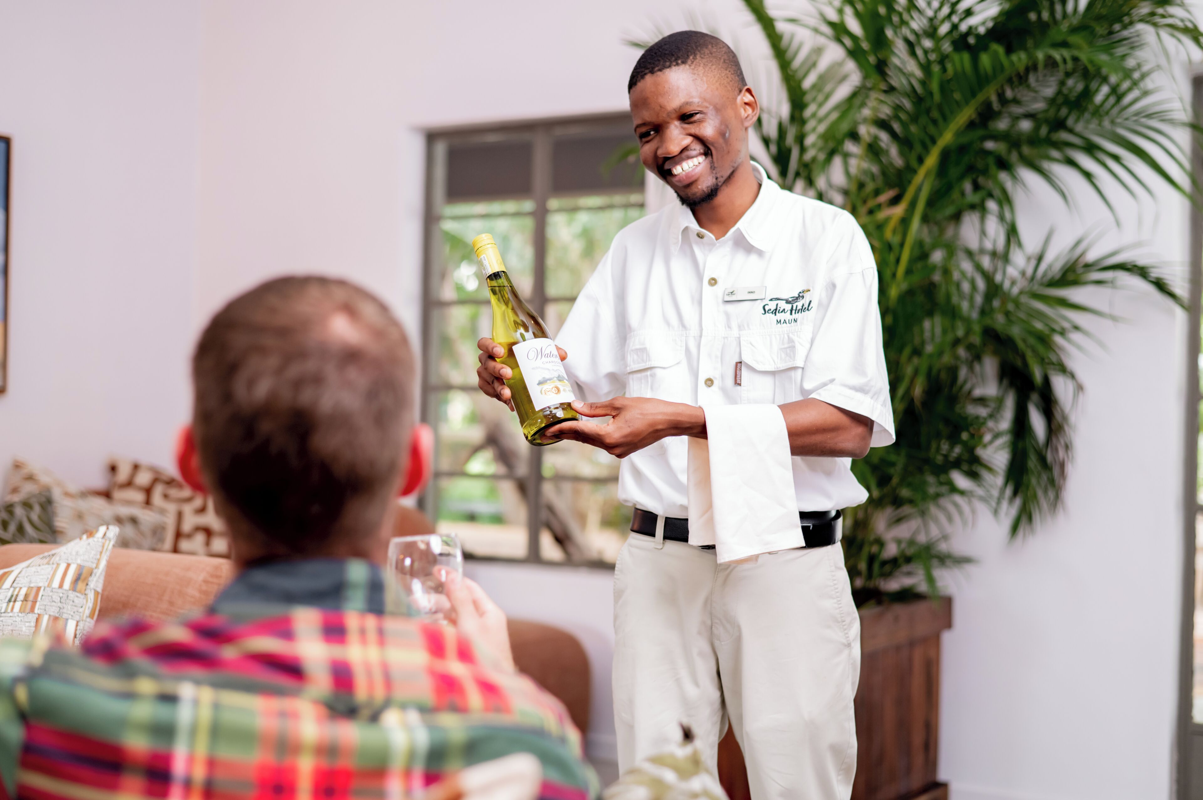 A person holding a wine bottle in a room with a window, a plant, and a couch.