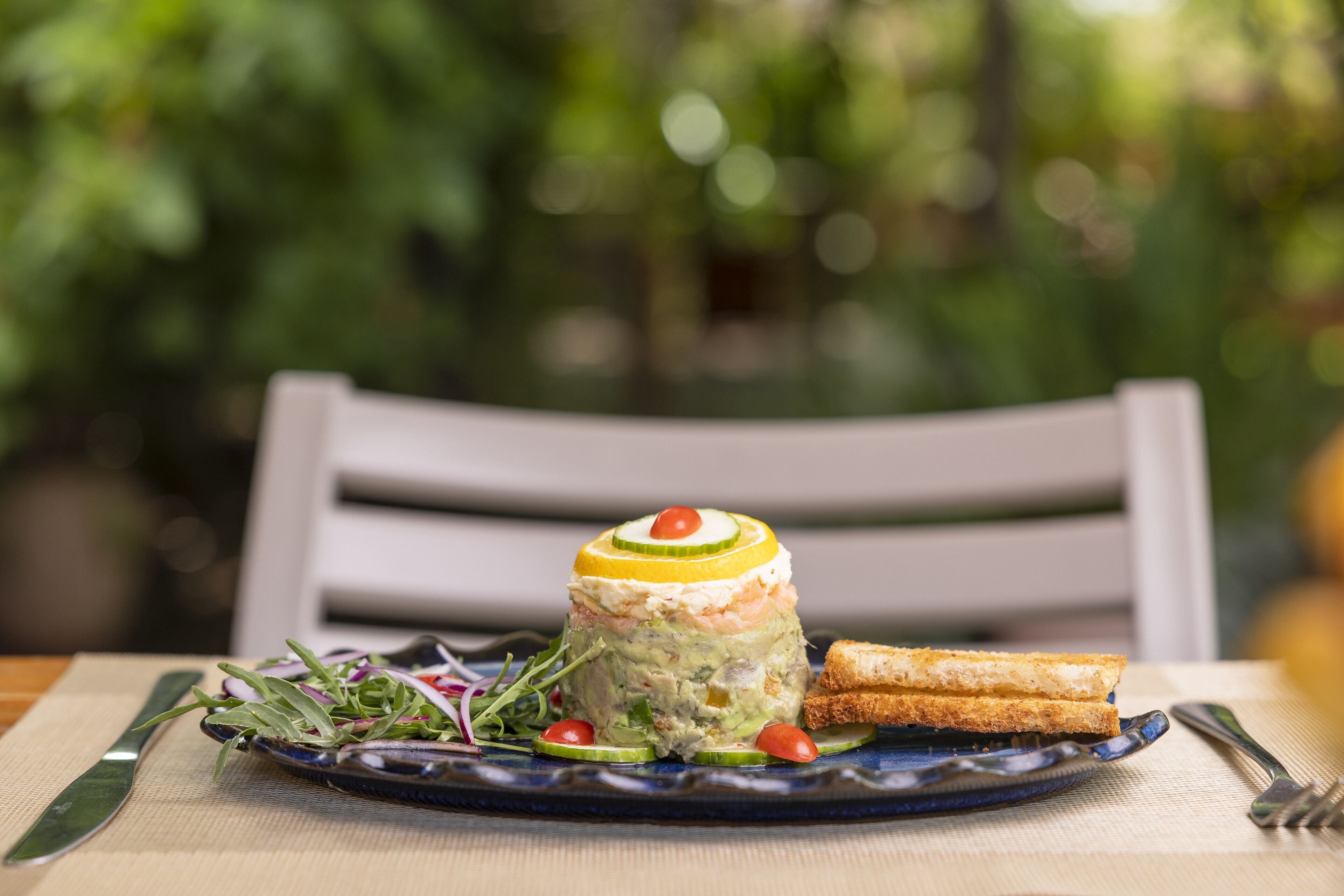 Un plato con una ensalada en capas, una rebanada de pan y un cuchillo y tenedor sobre una mesa, con una silla al fondo.